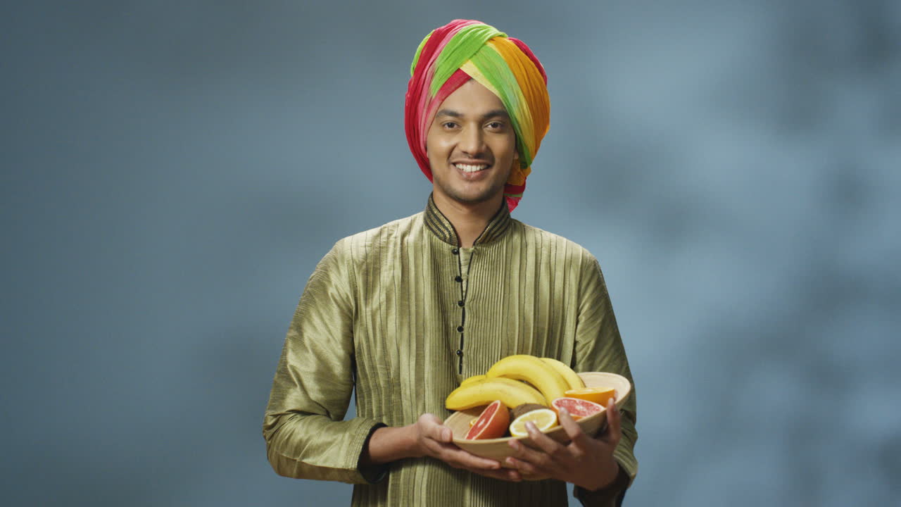 Young happy indian man in traditional clothes and turban smiling cheerfully at camera and holding plate with fruits