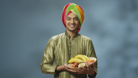 young happy indian man in traditional clothes and turban smiling cheerfully at camera and holding plate with fruits