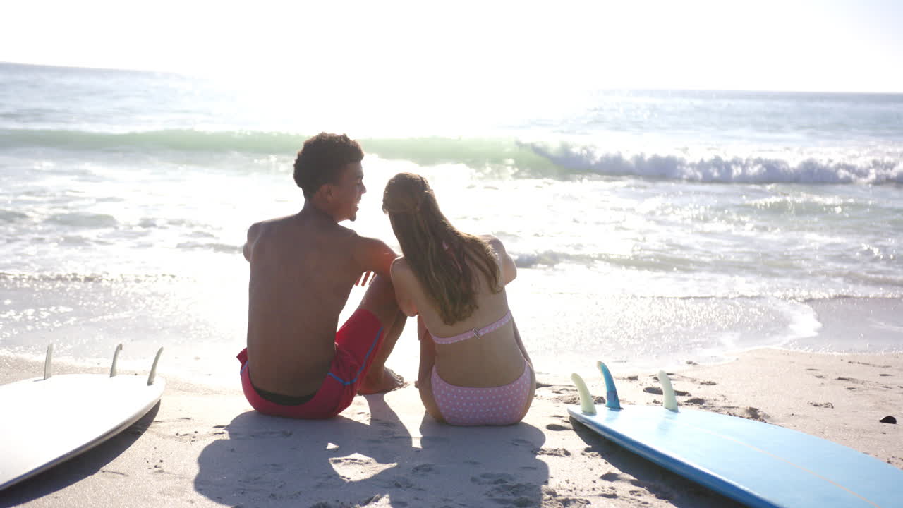 un hombre biracial y una mujer caucásica se sientan juntos en una playa soleada.