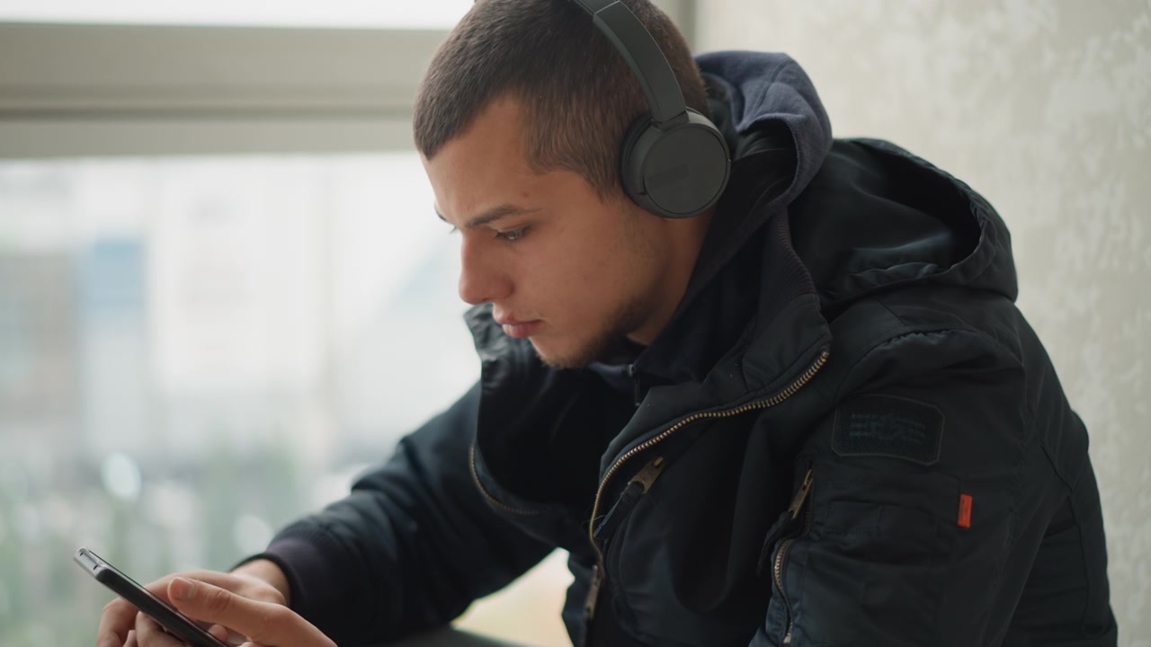 Close up of young man wearing black jacket and headphones holding smartphone in both hands while seated near window with blurred cityscape. Soft natural light illuminates scene emphasizing