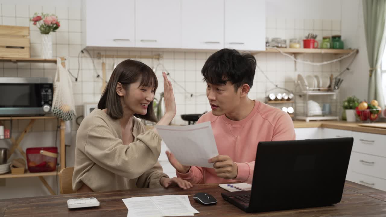 excited Asian spouses reading paper letter with good news in the dining room at home. they give high five and clap hands celebrating for getting tax refund