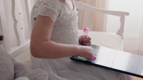 Hermosa-Niña-Usando-Una-Tableta-Dibujando-Imágenes-En-La-Pantalla-Táctil-Disfrutando-De-La-Creatividad-Sentada-En-Un-Banco-Con-Un-Osito-De-Peluche-Comiendo-Paleta-En-Casa