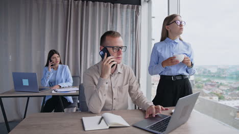 people working in office. employees using phones and laptops at their work. female colleague drinks coffee standing at window.