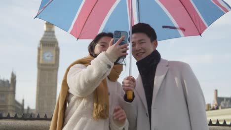 young asian couple on holiday posing for selfie in front of houses of parliament in london uk with umbrella