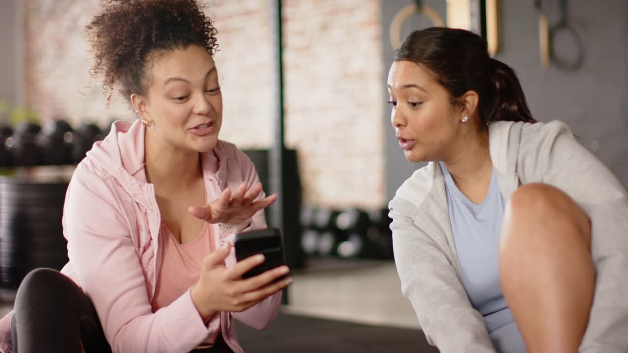Diverse female friends tilting smartphone toward friend and showing surprise on exercise mat in gym