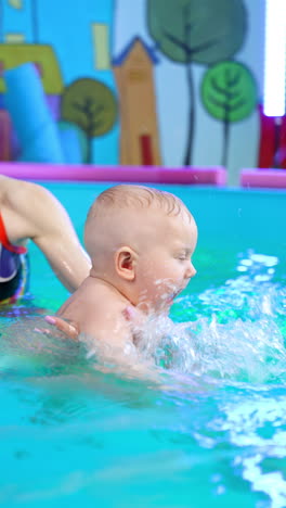 one-year-old toddler is being taught to swim. female coach moves the baby in water, happy child taps his hands cheerfully. vertical video.