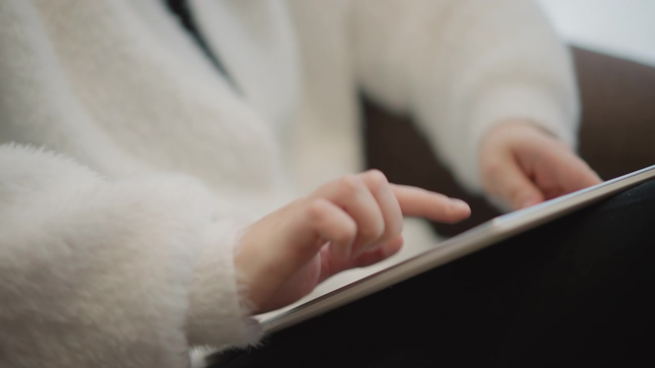 Low-angle close view shows hands in fluffy white sweater using horizontally held tablet indoors, with finger hovering, tapping, swiping, and changing screen in soft warm light, with hand, tablet