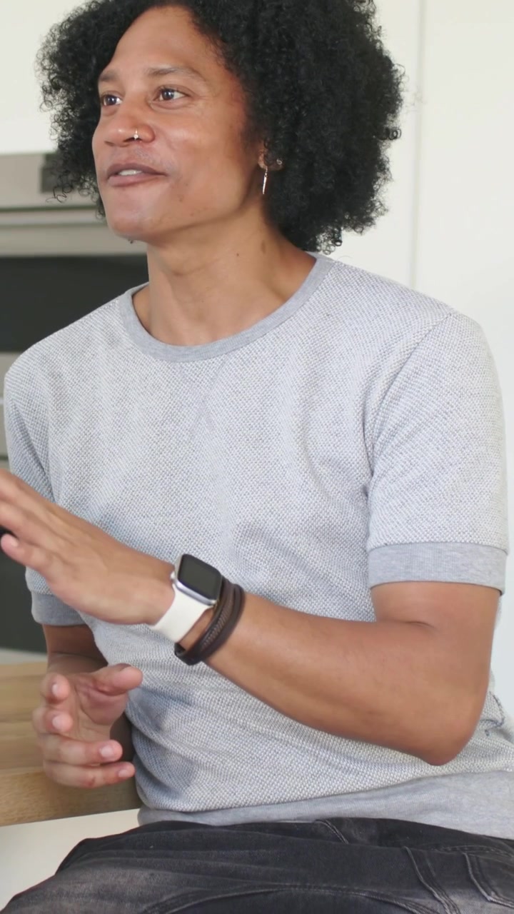 Vertical video: Responding to offscreen cue, man in grey tee speaking from kitchen, showing watch