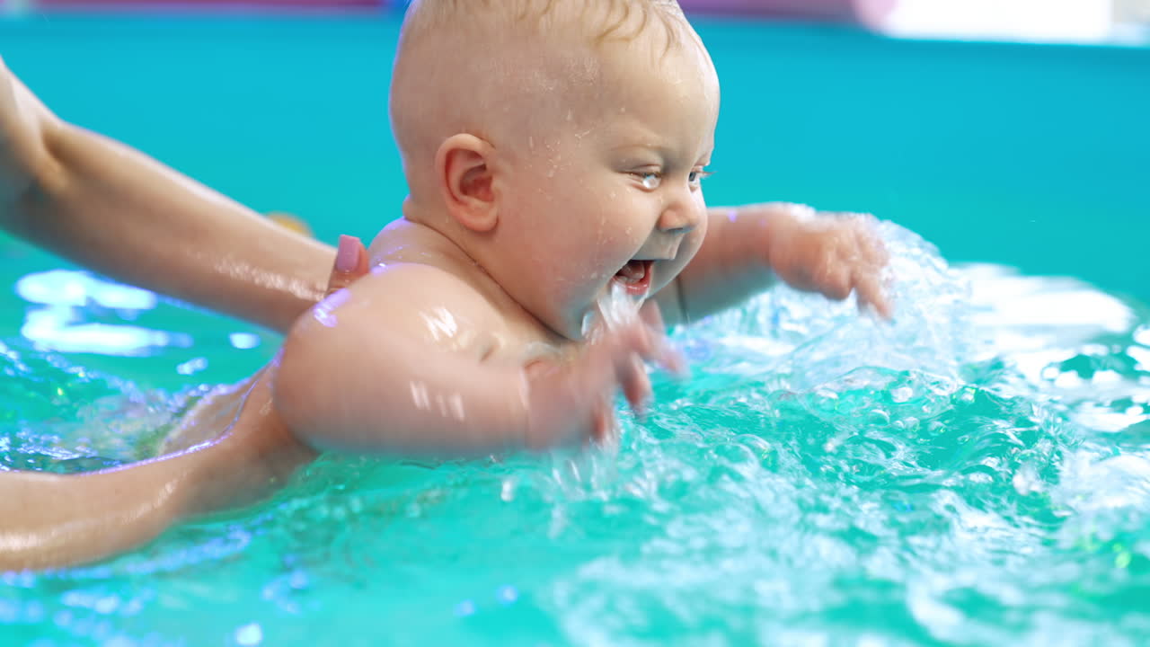 Cute smiling Caucasian baby in the swimming pool. Woman supports the child in water teaching him to swim.