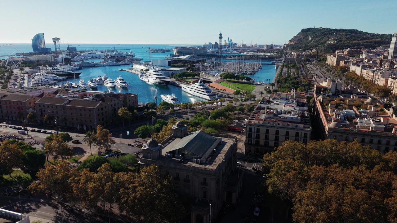 Drone panning over Barcelona harbor showing marina with yachts, surrounding buildings and clear blue water.