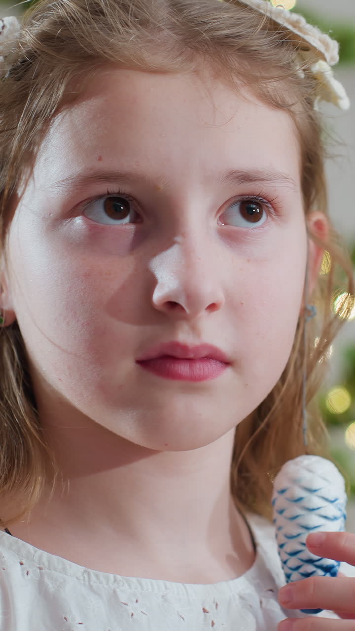 어린 백인 소녀가 크리스마스 트리를 장식하며 머리를 정리하고 솔방울 장식을 들고 있어요. 은은한 보케 조명과 함께 조용한 휴일의 집중, 실내 가족 풍경, 섬세한 드레스와 머리핀, 수줍음