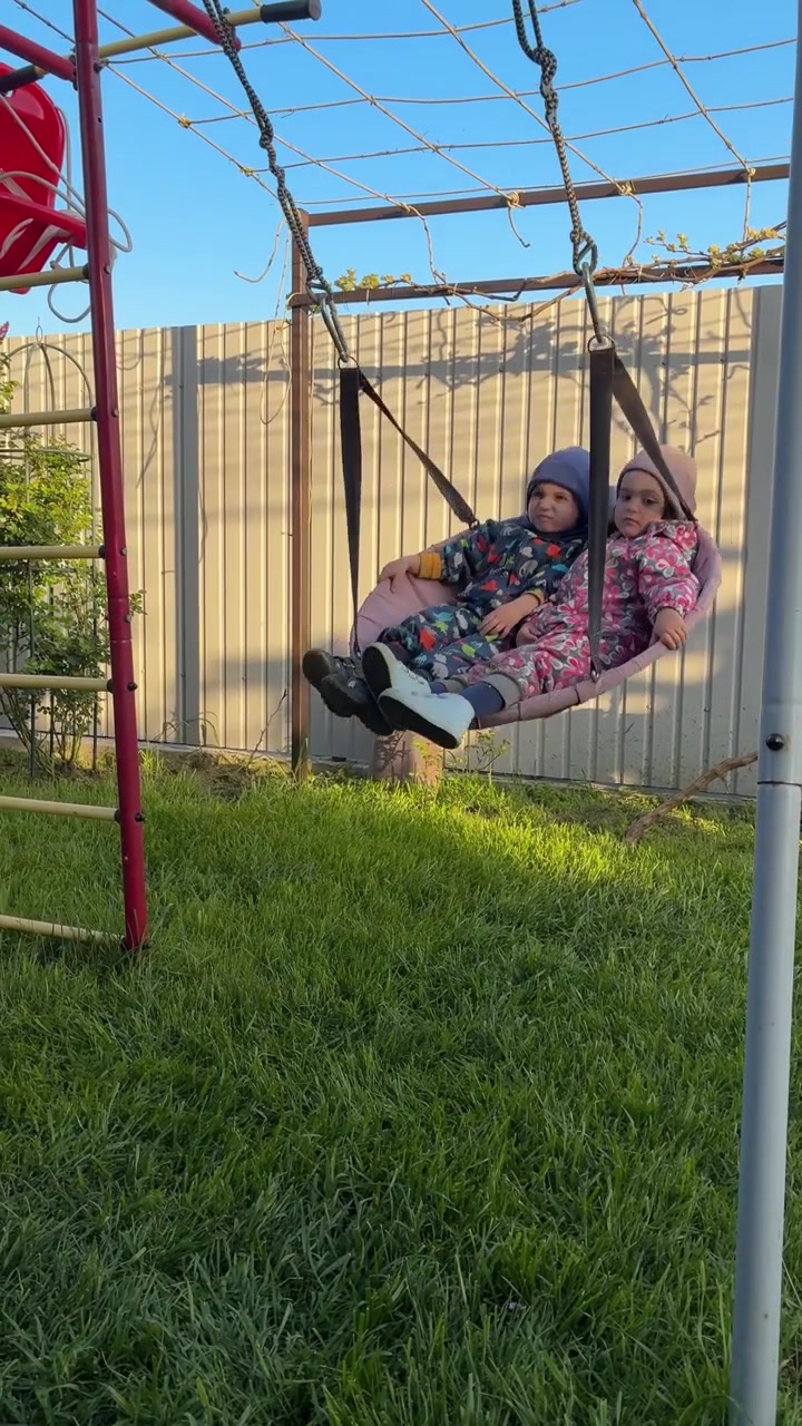 Due bambini piccoli con abiti colorati che dondolano avanti e indietro su un'altalena sospesa in un cortile soleggiato con erba verde e una recinzione