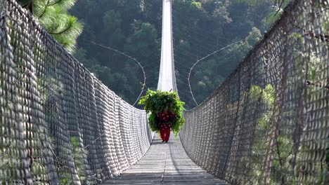 a nepali person carrying a heavy load and walking across a suspension bridge over a valley in nepal