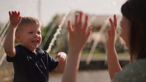 smiling boy waves hand splashing water on mother in park