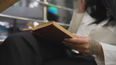 close up of college student seated in mall holding book on lap with tattoo hand visible, adjusting glasses while lights softly blink in background