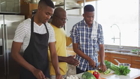 Afroamerikanischer-älterer-Vater-Und-Zwei-Erwachsene-Söhne-Stehen-In-Der-Küche,-Kochen-Abendessen-Und-Reden