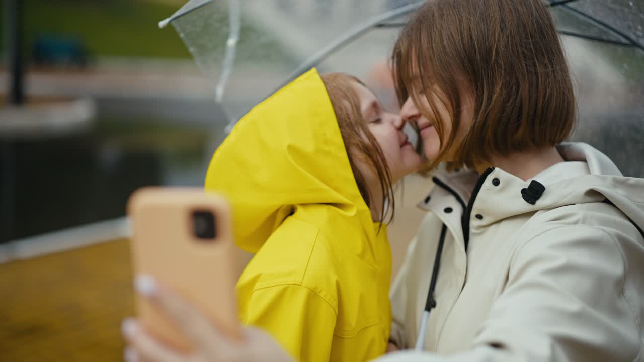 행복 한 금발 여자 가 방울 을 고 있는 길거리 에서 노란색 재 을 입은 십대 딸 과 셀피 를 찍고 있다