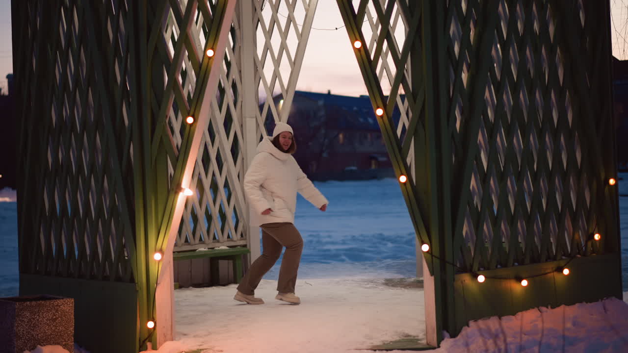 Choreographer practices rhythmic dance movements on snowy gazebo at dusk wearing white puffer coat beanie and headphones as gentle breeze tousles hair beneath bare tree limbs exuding winter joy
