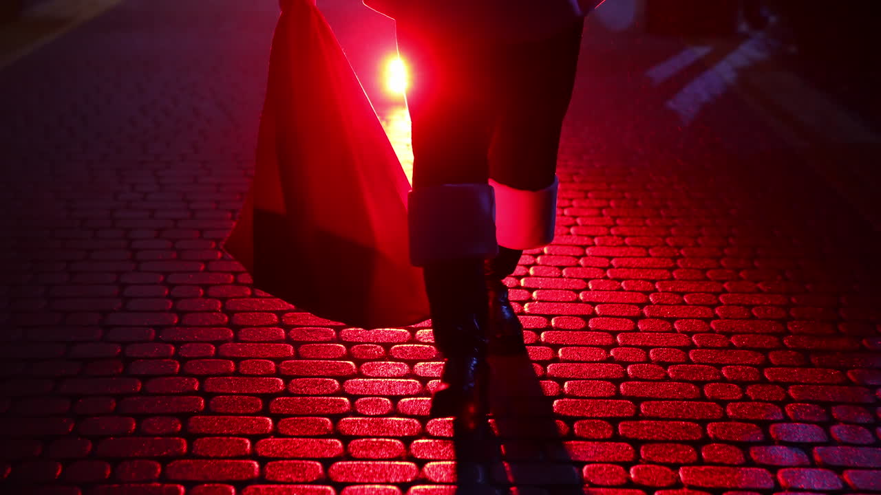 Legs of Santa Claus. Father Frost carrying presents at Christmas in red bag outdoors. Black boots of Santa walking on a pavement at dark on snow background. Red light.