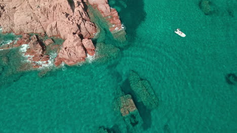 Vista-Estática-Desde-Arriba-Del-Mar-Transparente-Y-Cristalino-Donde-Las-Olas-Chocan-Contra-Las-Rocas-Rojas-En-Un-Bote-Con-Turistas-Nadando-Bajo-El-Sol-En-Cerdeña,-Italia---Toma-Aérea