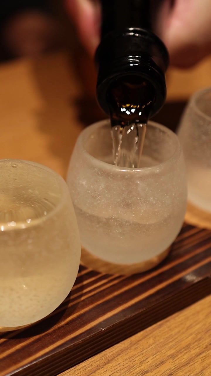 Tangkapan vertikal yang menunjukkan sake bening dituangkan dari botol gelap ke dalam cangkir kaca buram.