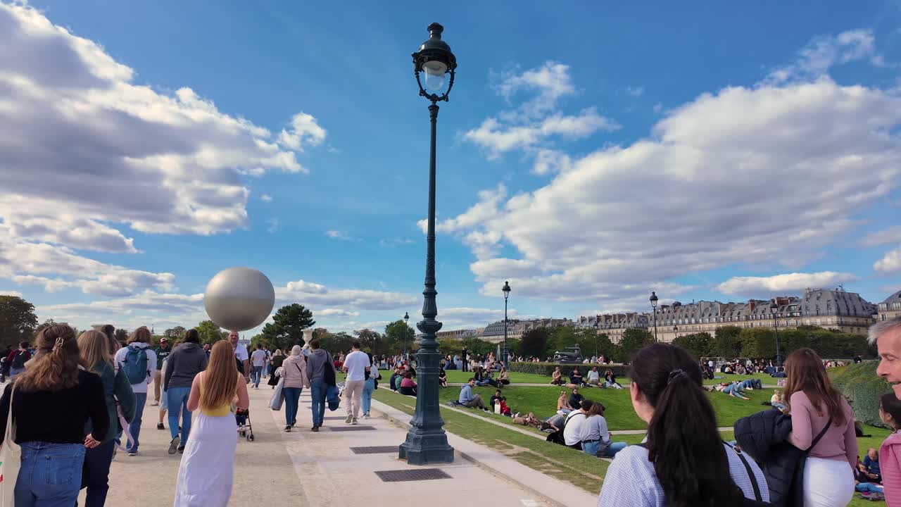 Crowds of people enjoying a sunny day in the Champ de Mars park in Paris with a large sphere sculpture in the background