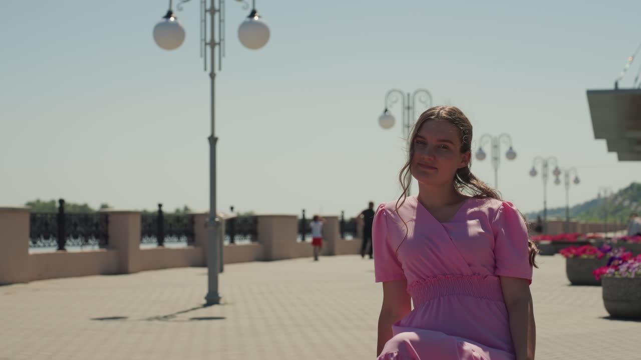 Mulher de vestido rosa sentada no calçadão, canteiros de flores vibrantes em primeiro plano, passarela com postes de luz, pose editorial com sorriso gentil, pétalas iluminadas pelo sol, tons pastel, carisma de modelo relaxada, viagem