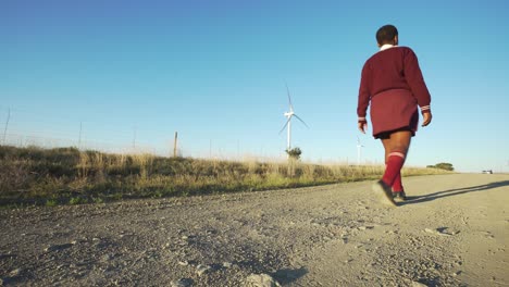 niña de la escuela caminando por un camino de grava junto a la turbina eólica, ángulo bajo