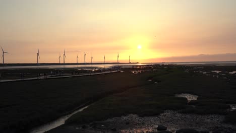 Spectacular-orange-sunset-landscape-shot-of-tourists-strolling-on-wooden-walkway-platform-at-beautiful-Gaomei-wetlands-preservation-area-with-wind-power-plant-in-the-background-in-Taichung,-Taiwan