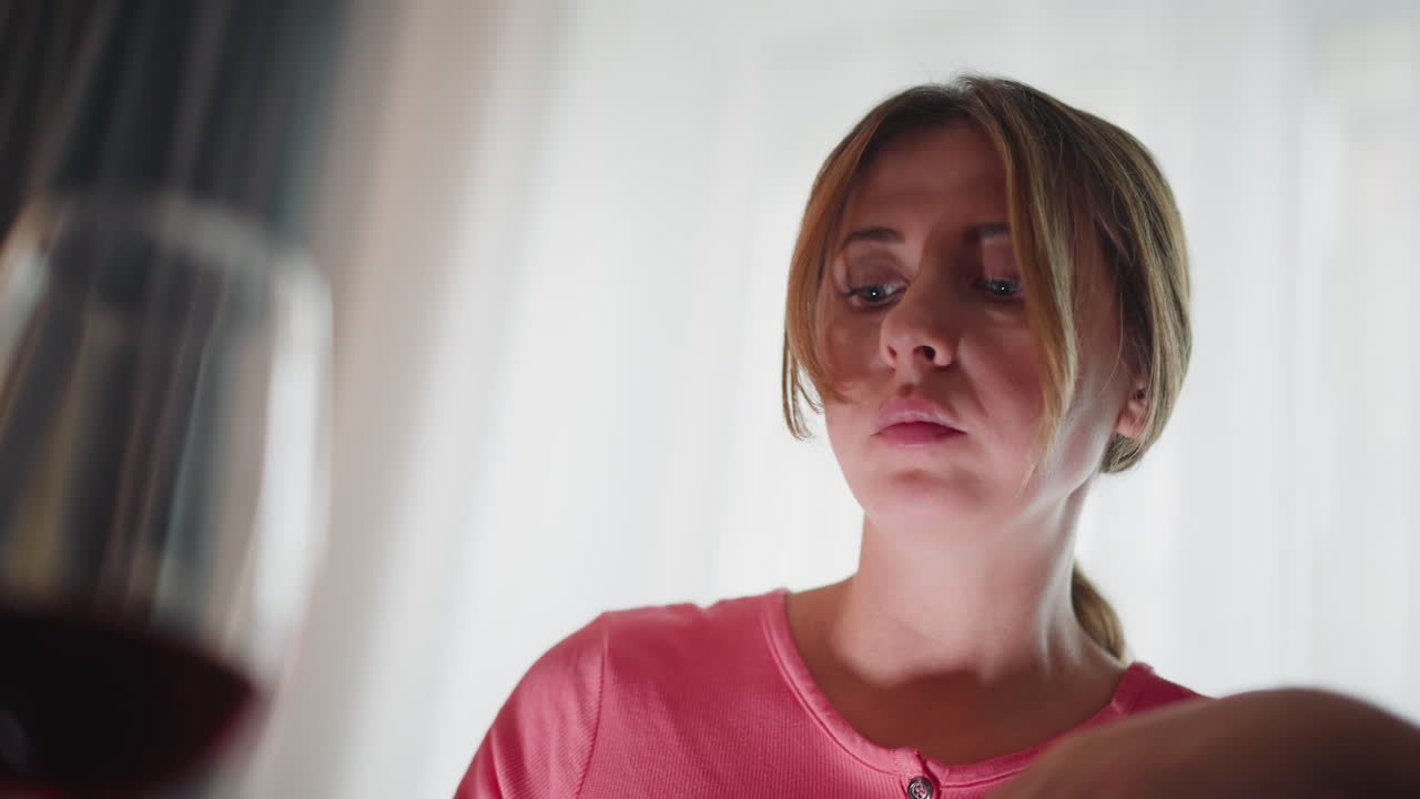 Student in pink top eating dinner using chopstick with sunlight highlighting neck while seated indoors with soft curtain-filtered natural light and glass of red wine visible in foreground
