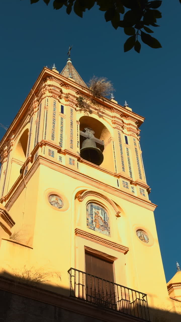 Campanile illuminato dal sole incorniciato da foglie, facciata calda e dorata contro un cielo blu intenso, prospettiva dal basso, ornato.