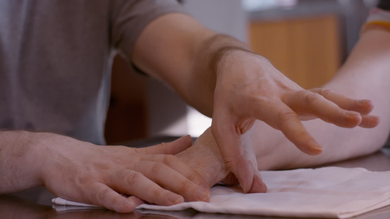 An adult man caregiver assists an elderly woman stroke survivor by gently stimulating and mobilizing her hemiplegic right thumb during a hand therapy session at home kitchen table, on cloth pad.
