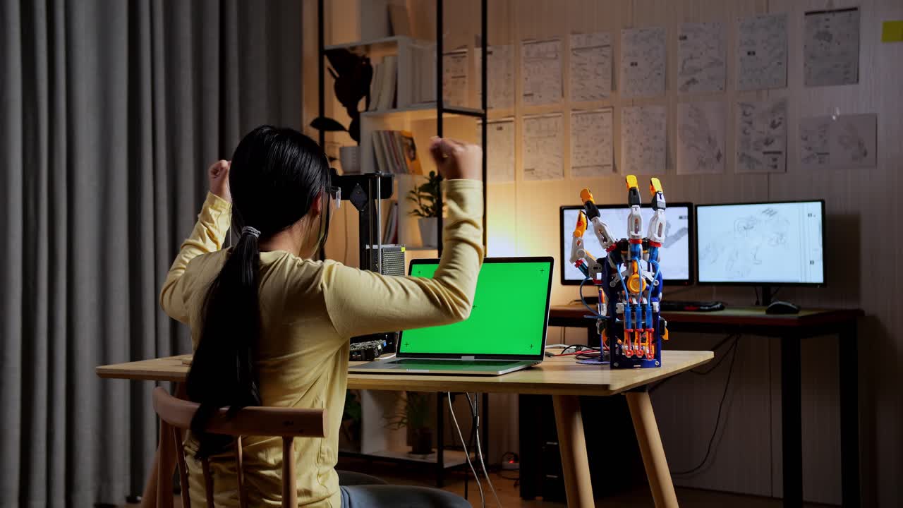 십대 아시아 소녀가 성공을 축하하는 뒷면: 집에서 녹색 화면 모형으로 노트북에 사이보그 손을 설계하는 것