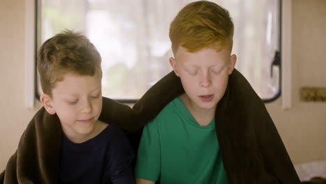 two little brothers with blanket on their shoulders sitting on bed in a campervan and reading a book