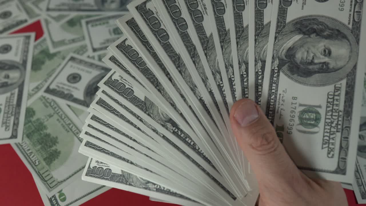 toma cerrada de una mano masculina que se mueve con una pila de cientos de dólares estadounidenses frente a una mesa roja llena de dinero