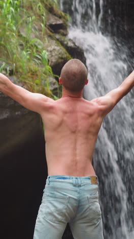 man tourist raises up hands enjoying power of wild nature near waterfall with large rocks at highland on summer day backside view slow motion