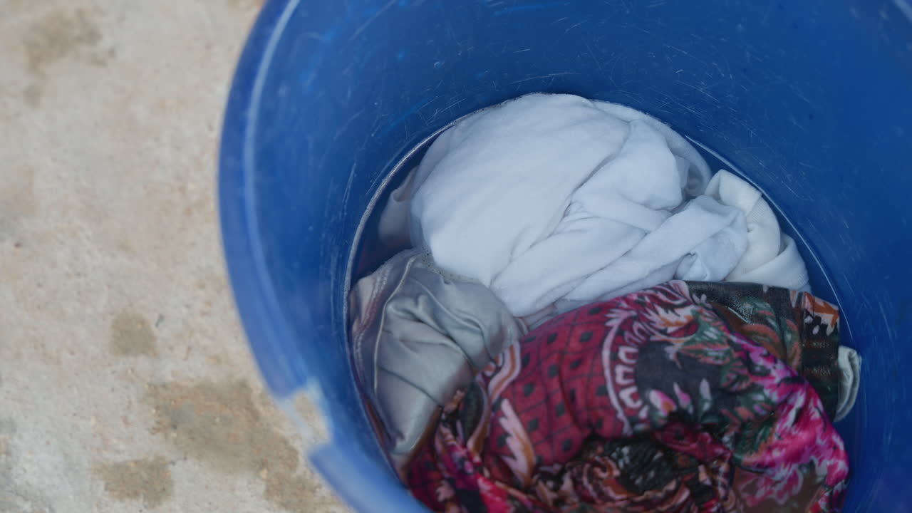 Soaked clothes in blue bucket, outdoor house chore on concrete floor, soapy water soaking colorful towel and patterned garment, fabric texture and wet folds, quiet domestic routine and utility scene