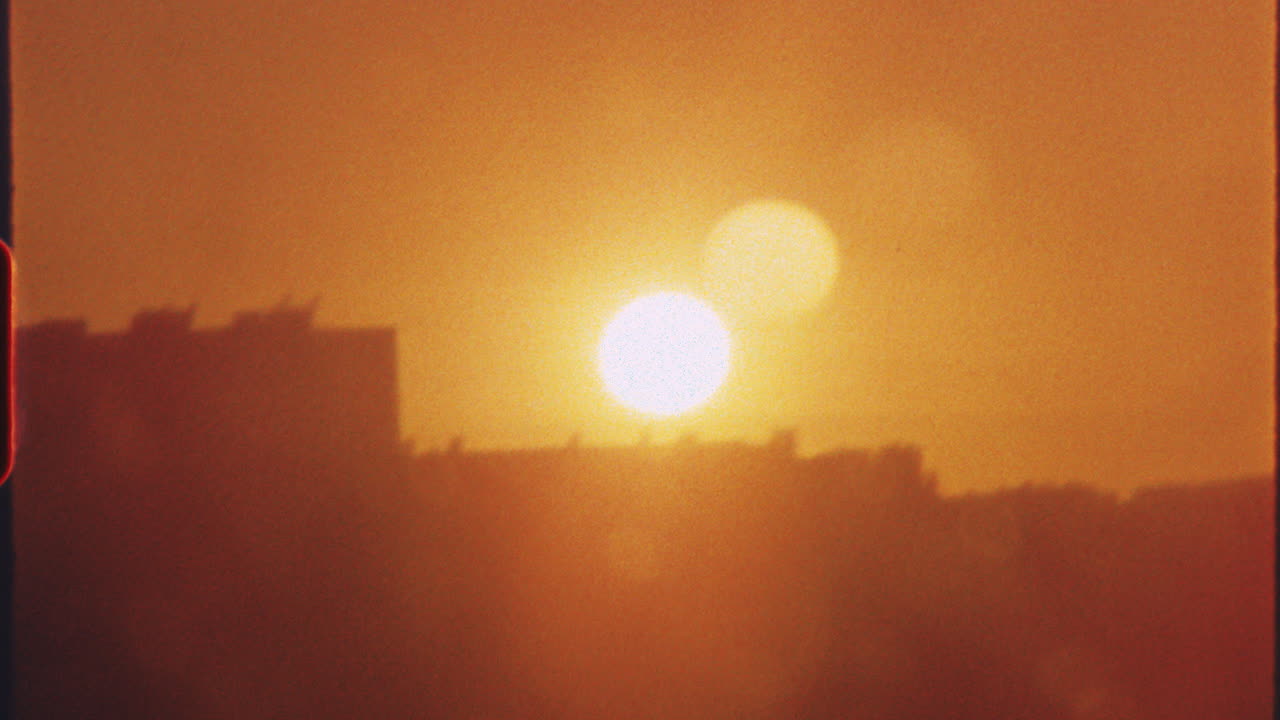 8 mm film - dramatic orange sun descending behind dark urban silhouettes, revealing warm golden hour light across architectural cityscape with gradual color transitions and tranquil atmospheric depth