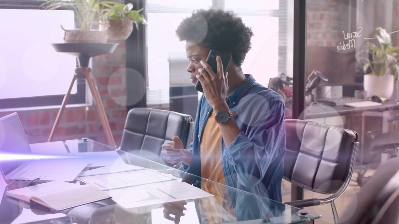 Man at glass-top desk answering phone call, scanning charts while lens-flare overlaying business