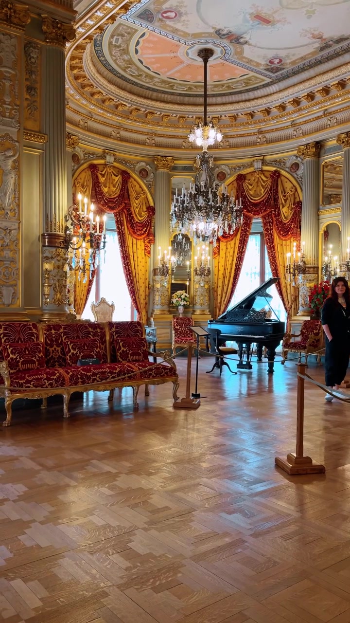 Visitatore che cammina attraverso una grande sala storica con decorazioni ornate. Vista posteriore di una persona in una sala di museo con colonne dorate, drappeggi rossi, un pianoforte e un soffitto decorato