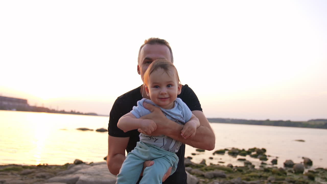 Loving father with cute little toddler in his arms approaching camera. Adorable baby boy is laughing cheerfully. River at backdrop.