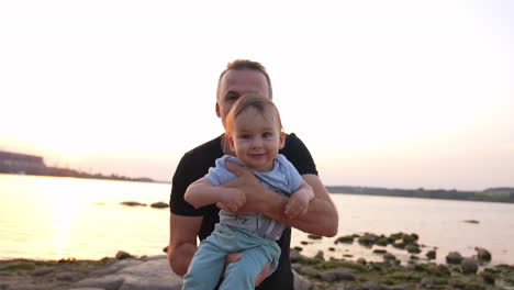 loving father with cute little toddler in his arms approaching camera. adorable baby boy is laughing cheerfully. river at backdrop.