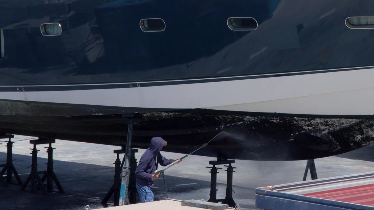 Caucasian worker uses high-pressure water to clean a large yacht hull in a shipyard.