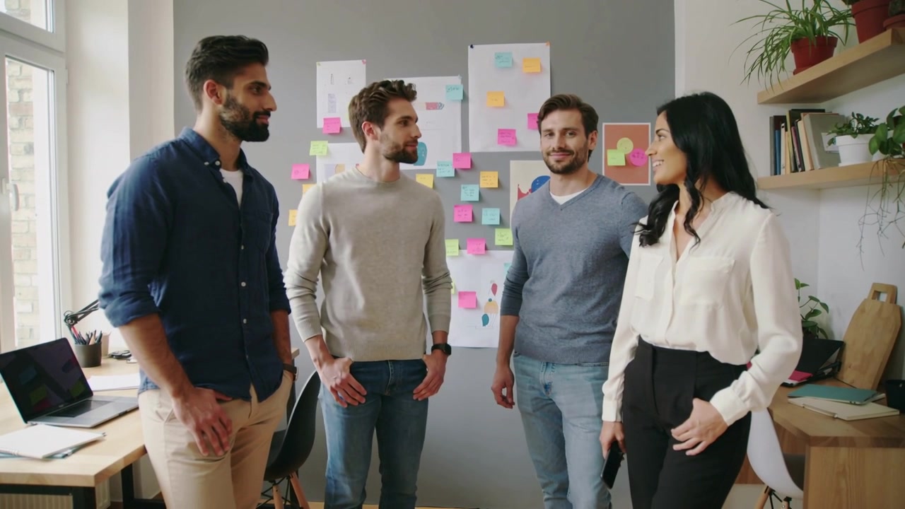 Fire kolleger i business-casual tøj reagerer på manden til venstre, der taler, mens de samarbejder om noter på kontoret.