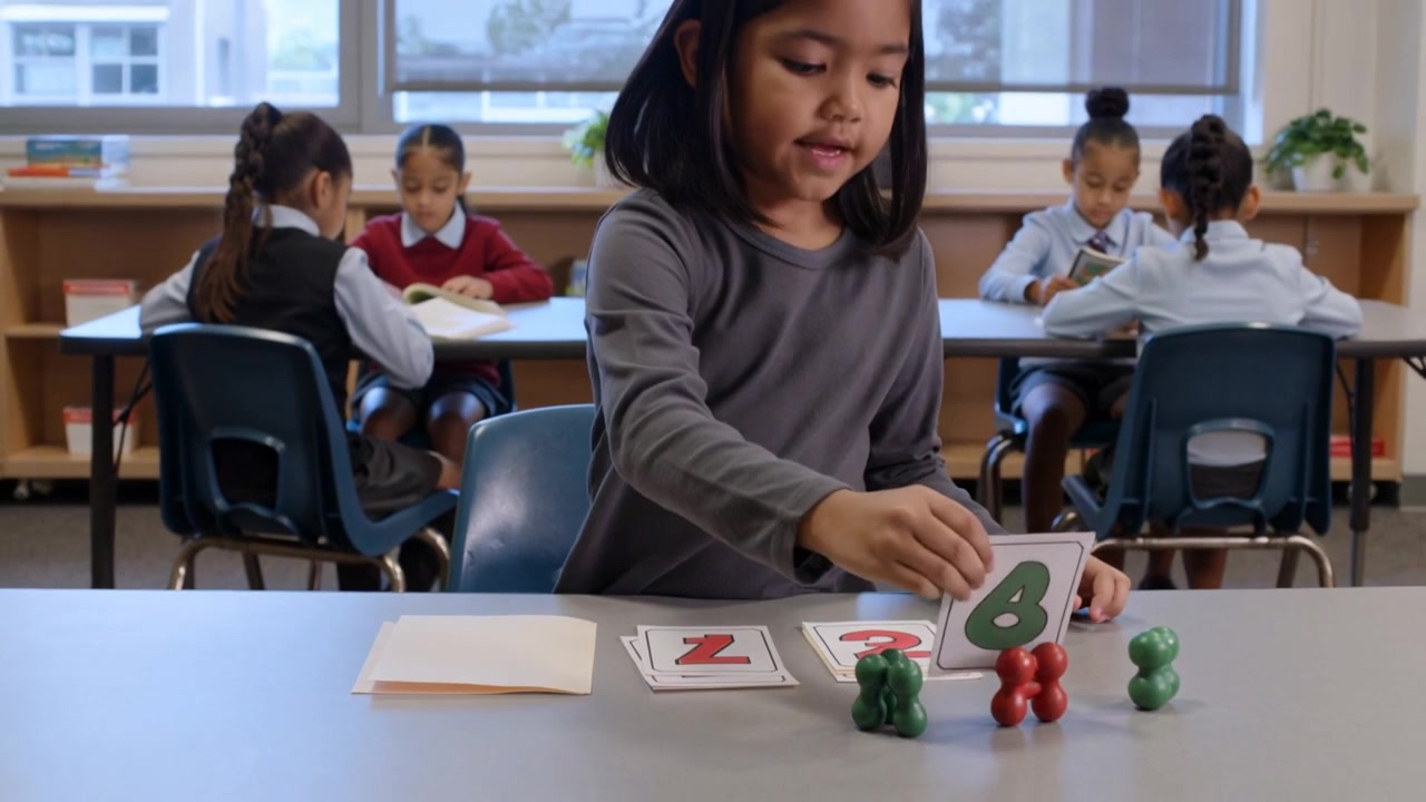 Picking cards student in gray top at classroom table placing green 8 on folder, matching counters