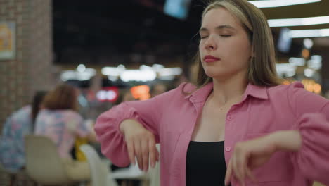 lady in pink closes her laptop in a busy restaurant, looking bored, she tilts her head from side to side, stretches, and clasps her hands together as people are seated and chatting around her