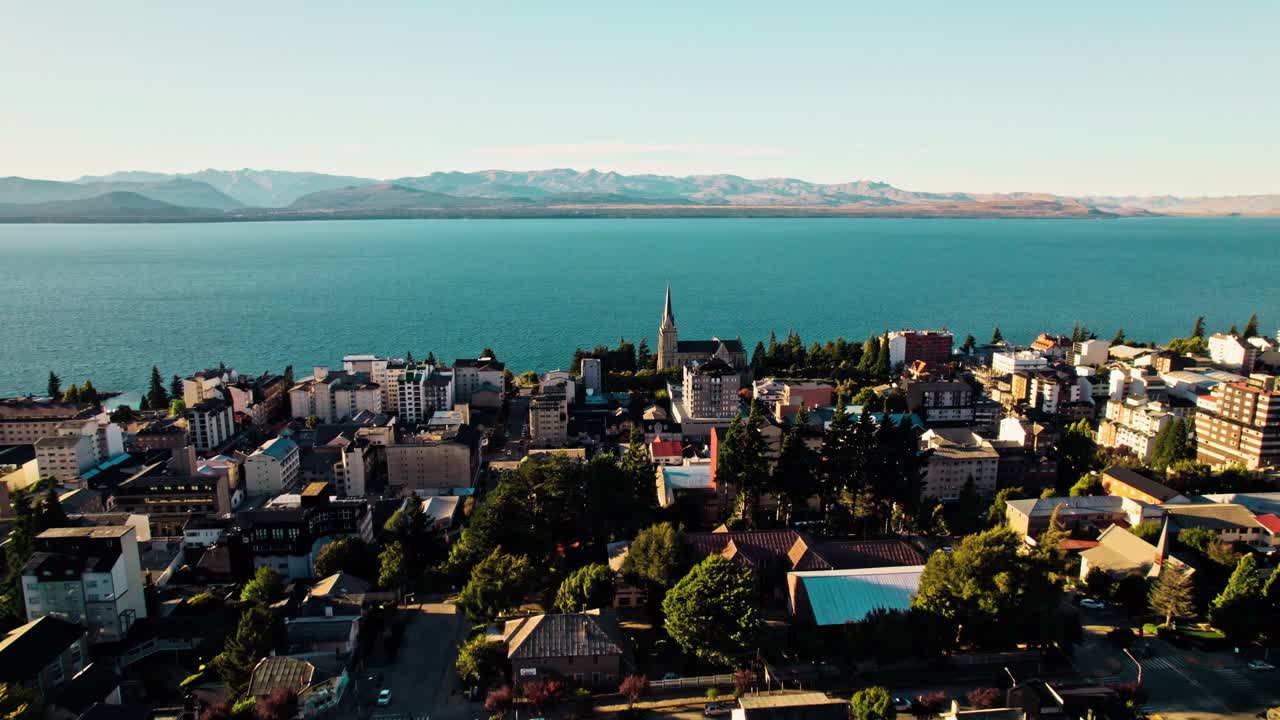 Wide drone shot of San Carlos de Bariloche urban center, highlighting the historic Cathedral near the lake, with a vast backdrop of the Nahuel Huapi and the arid Patagonian steppe beyond