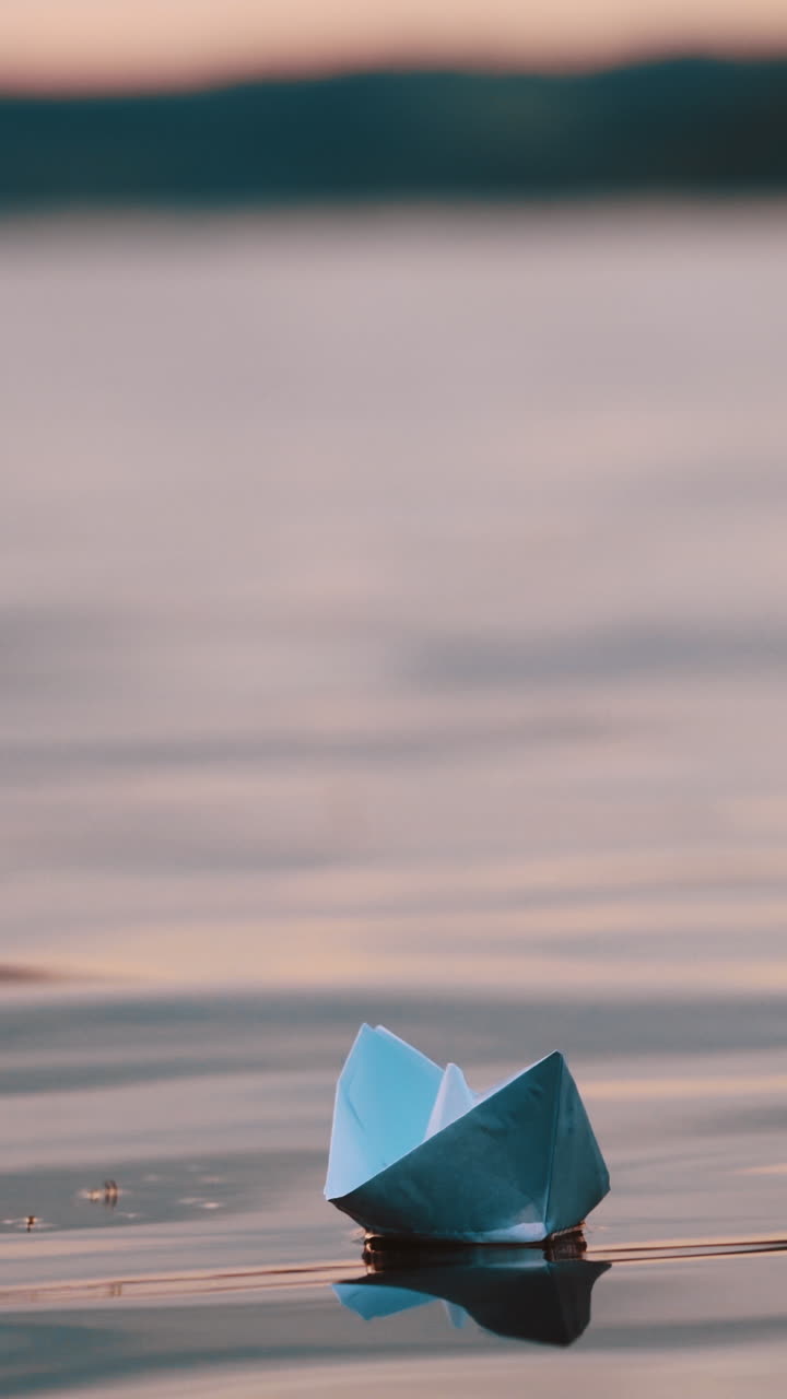 Blue homemade boat on water on blurred evening background. Paper little ship floating alone in calm river at sunset. Vertical video
