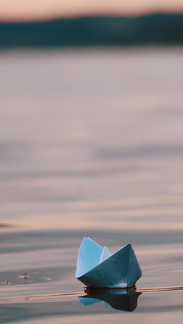 blue homemade boat on water on blurred evening background. paper little ship floating alone in calm river at sunset. vertical video