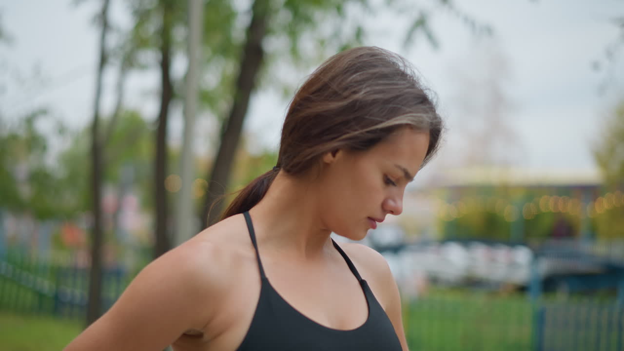 Sporty girl in neck top stretching her neck slowly in park, blurred background featuring decorated lights, trees, and park elements, capturing relaxation, outdoor wellness, and neck exercises